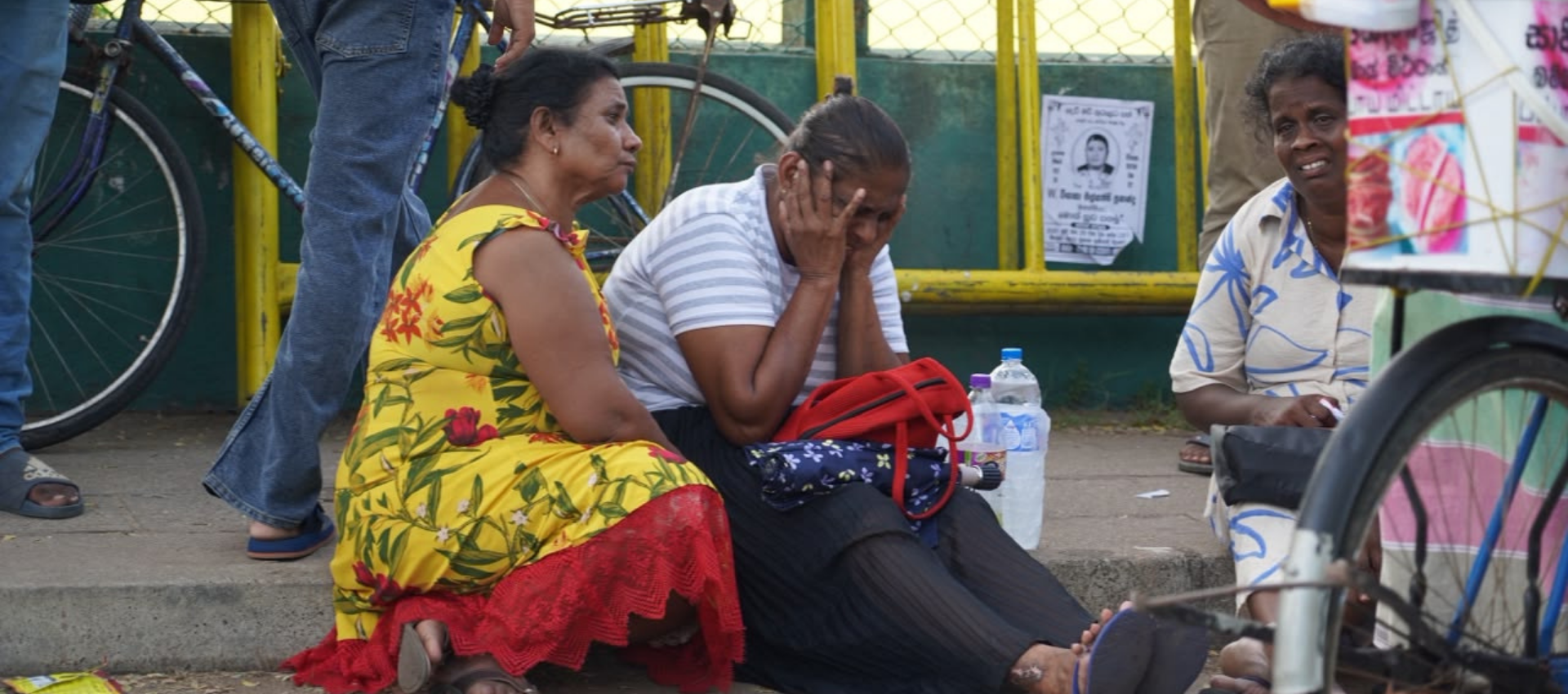 The image shows three women sitting down on a road. The lady sitting in the middle is seen breaking down with her hands covering her face, while other women appeared to be emotional as well.