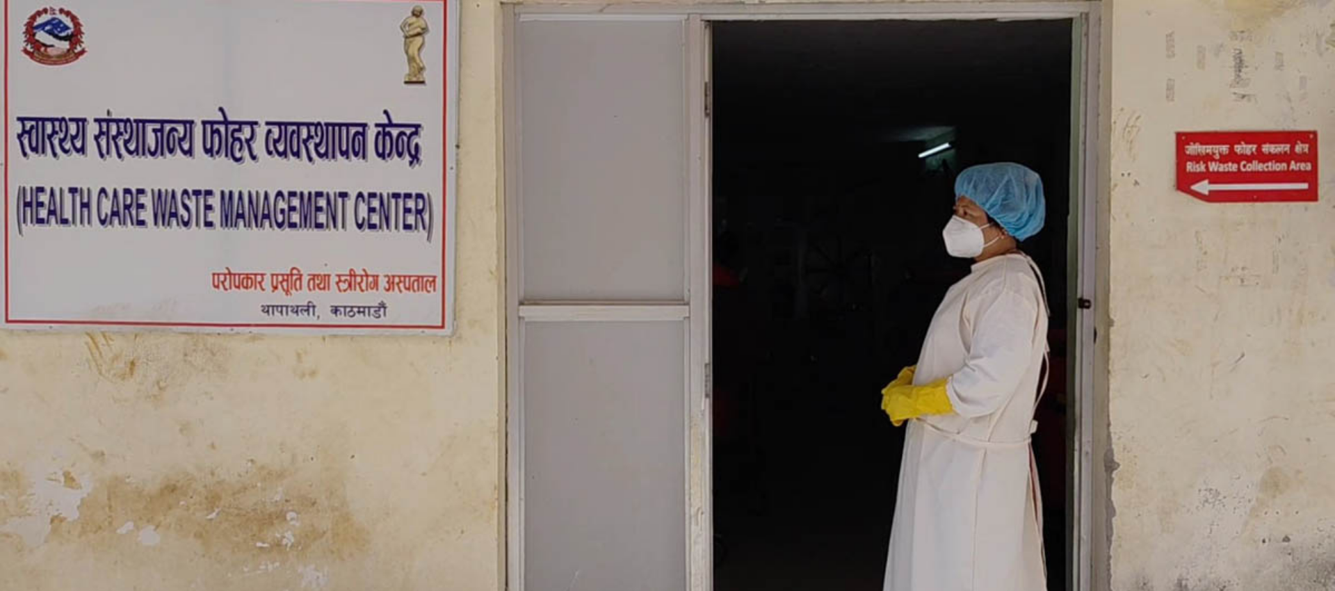 The image shows a woman standing outside the door of a healthcare waste management center in Kathmandu, Nepal. The woman can be seen wearing protective clothing.