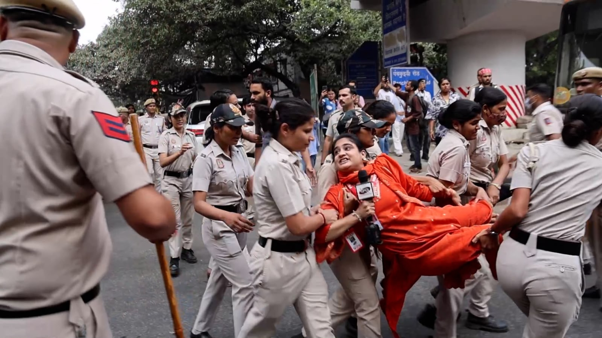 Police personnel carrying a female journalist with mic in her hand as bystanders watch the scene.